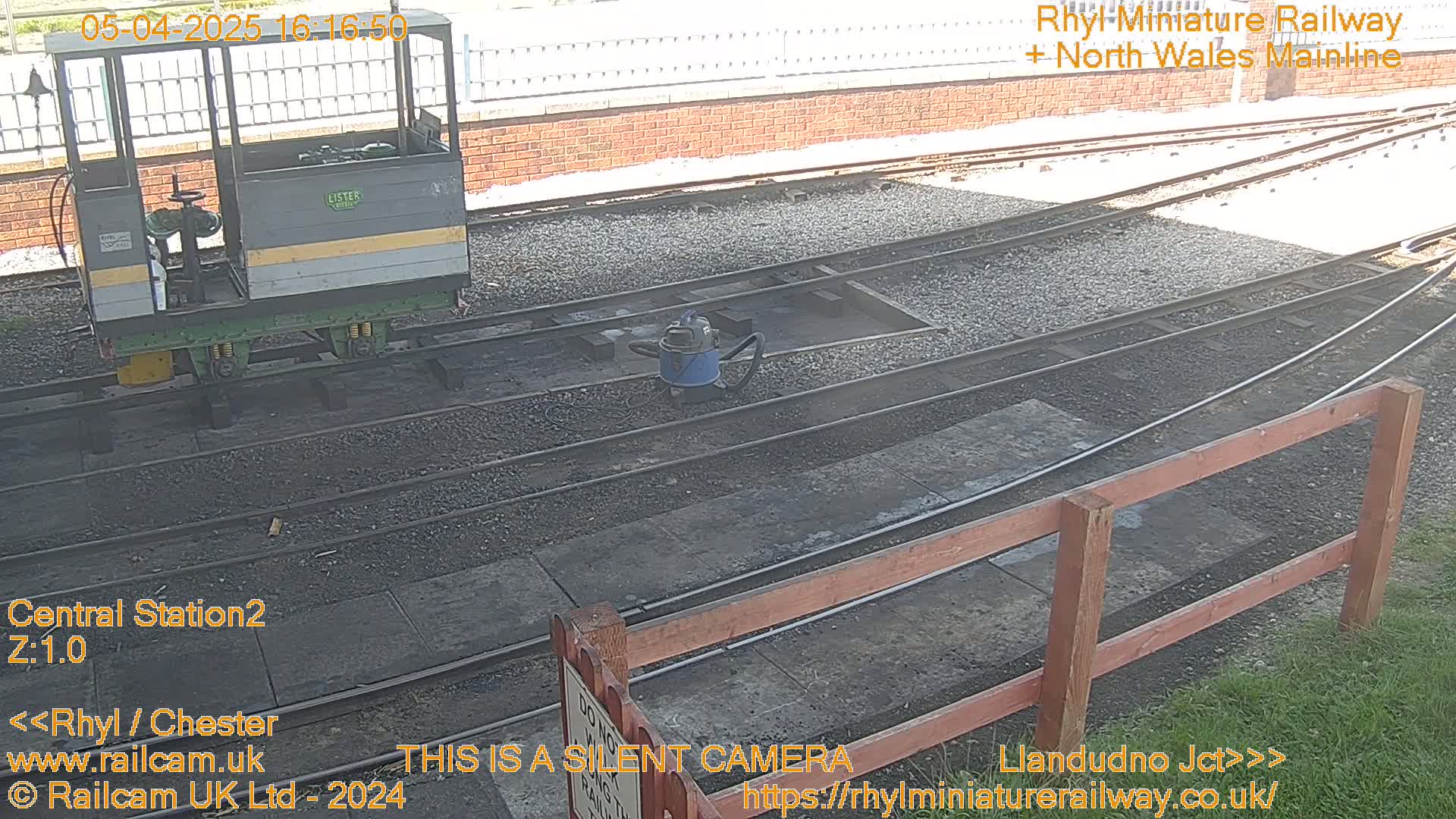 A small railway car sits near a section of railway tracks on a sunny day, with a vacuum cleaner visible nearby.