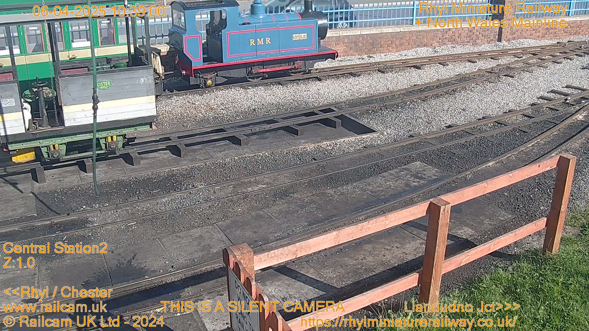 Two miniature trains are parked on a sunny day next to a railway track and wooden fence.