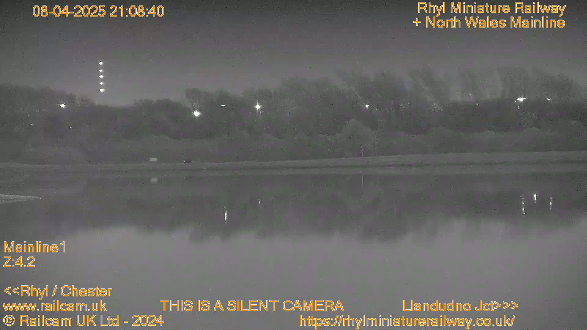 A nighttime, low-light image shows a body of still water reflecting lights from the far bank, where trees and distant lights are visible.