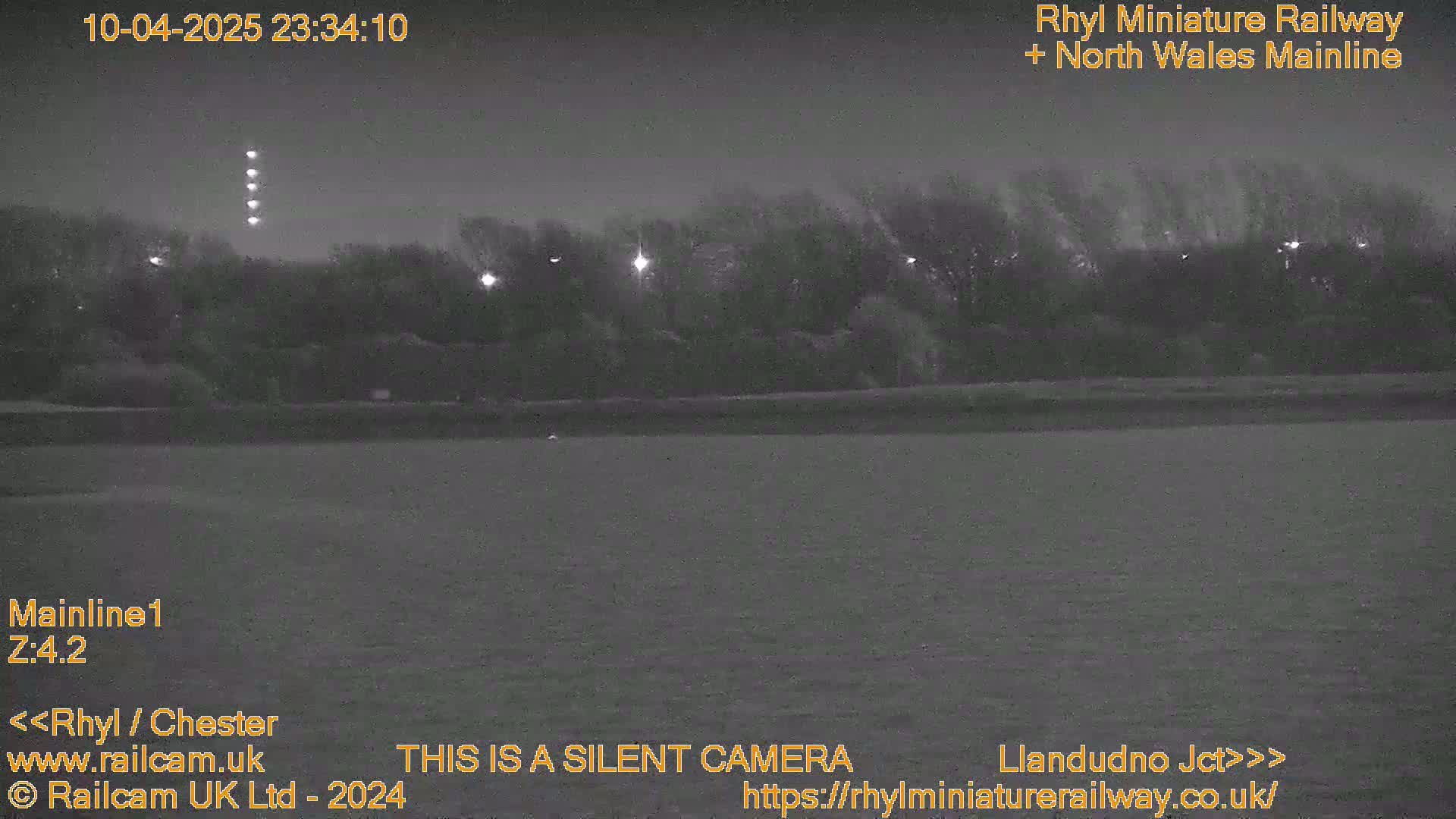A nighttime, low-light view of a flat, grassy field with trees and lights in the background.