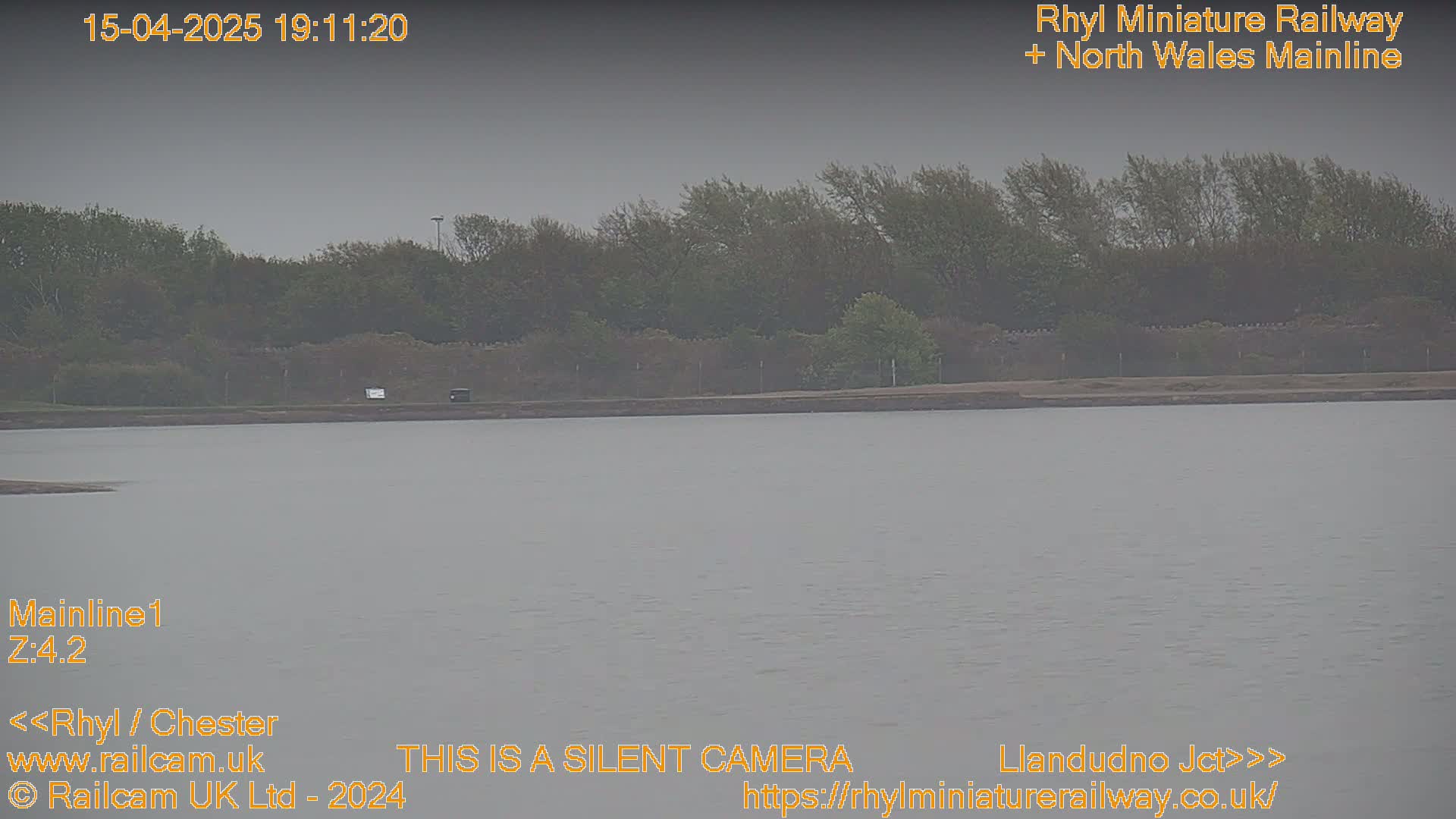 A body of still water is seen under an overcast sky, with a line of trees and a low wall visible in the background.