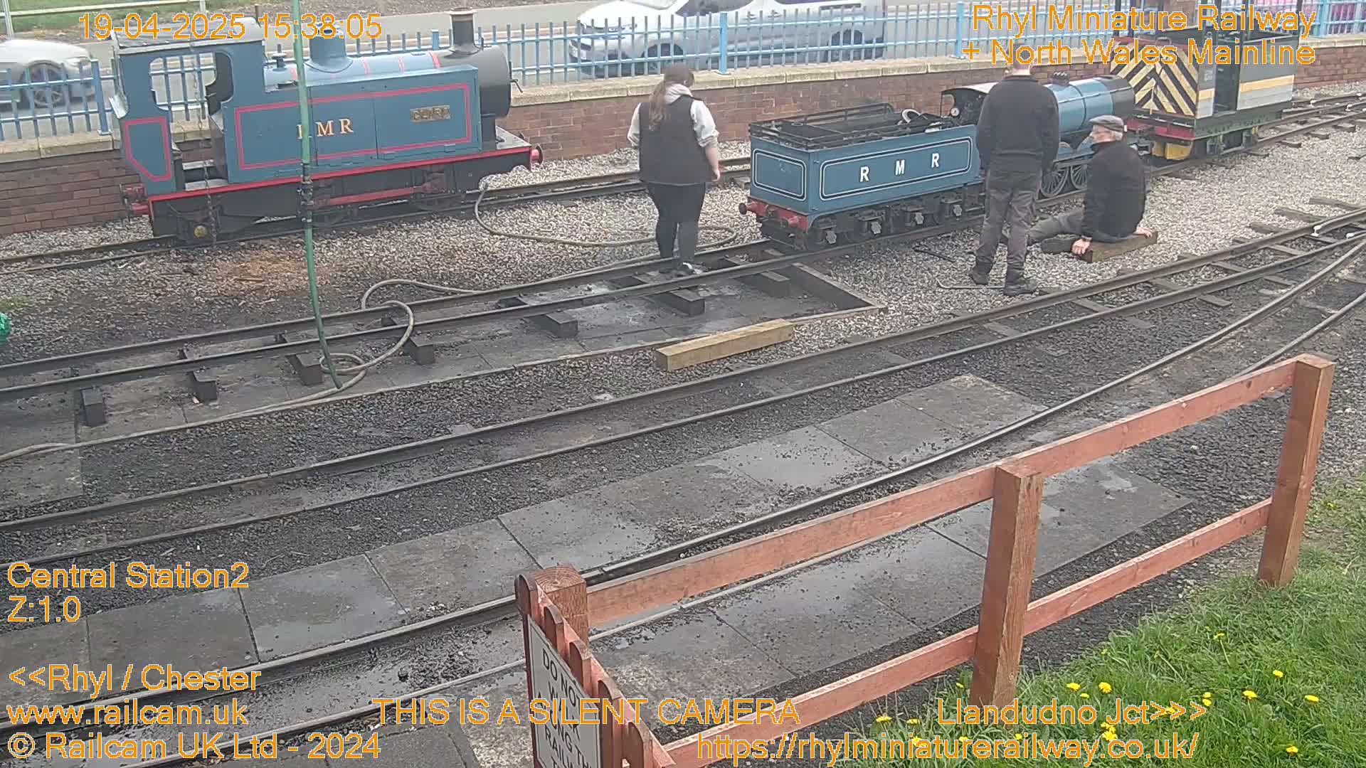 Two miniature train engines are parked on railway tracks next to several people, on a slightly damp day.