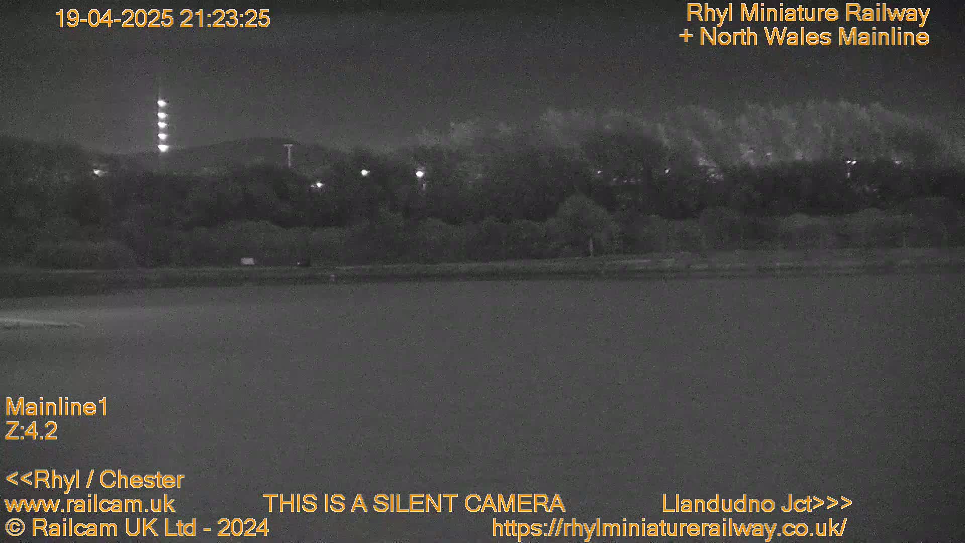 A nighttime, low-light image shows a dark body of water in the foreground, with a line of trees and lights behind it.