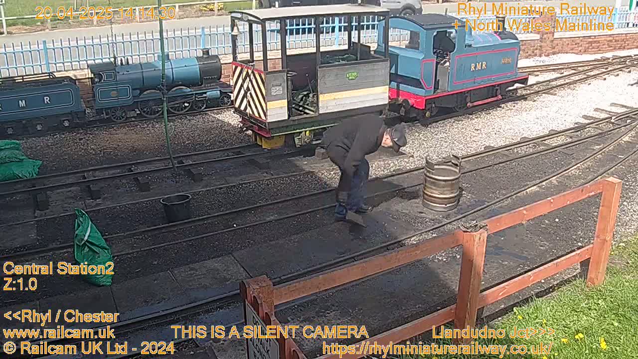 A person bends over near train tracks next to several small model trains and a metal barrel on a sunny day.