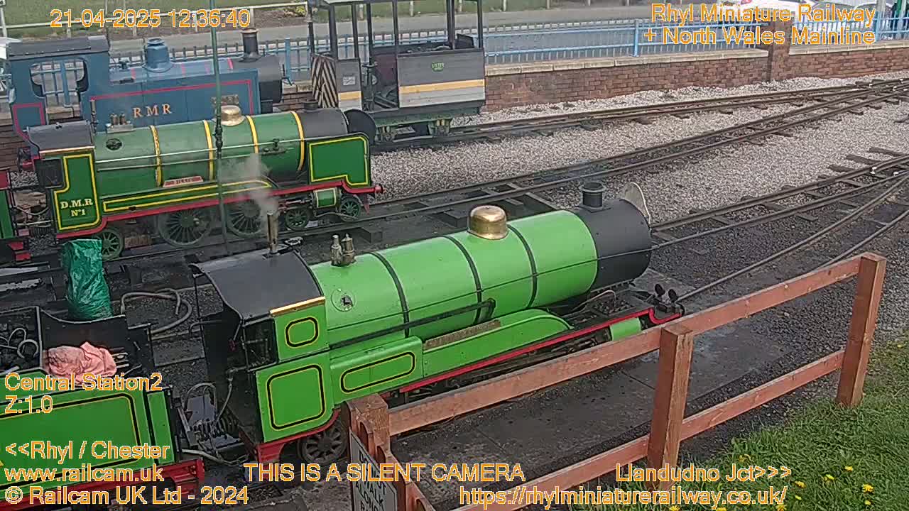 Two green miniature locomotives sit on railway tracks in an outdoor setting on a partly cloudy day.