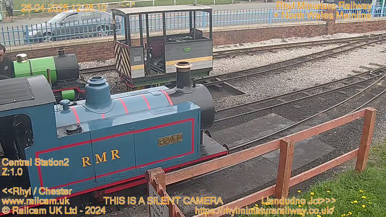 Three miniature railway engines are visible on a track, one of which is blue, in a sunny outdoor setting.