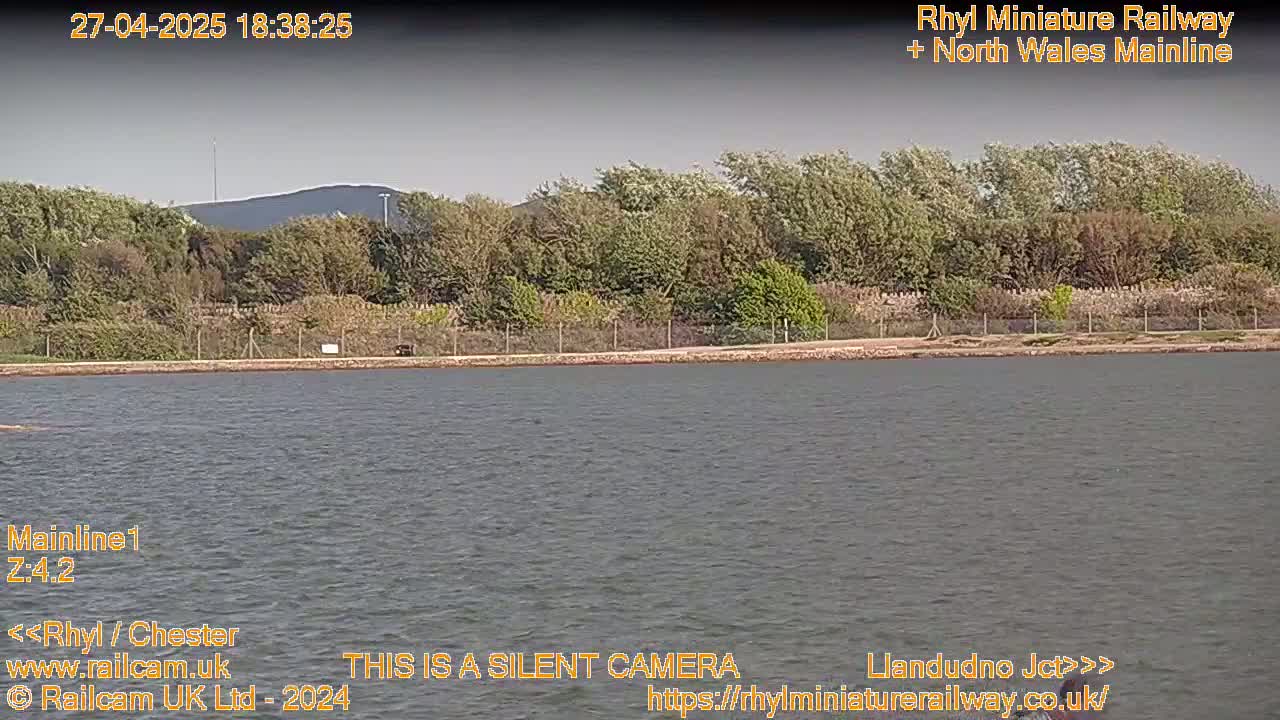A calm body of water sits before a line of trees and a distant hill under a mostly clear sky.