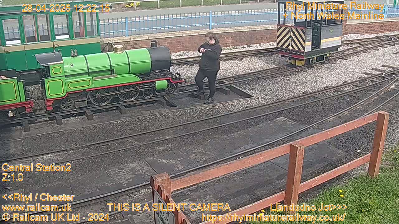 A person stands near a green miniature train on a railway track, and another small train is visible in the background on a sunny day.