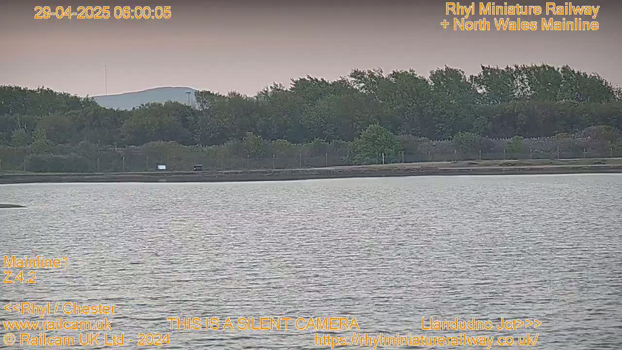 A calm body of water stretches out before a line of trees and a distant hill under a dusky, overcast sky.