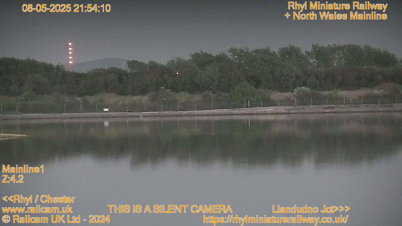A calm body of water reflects a dark, tree-lined shore under a dusky sky, illuminated by several bright lights in the distance.