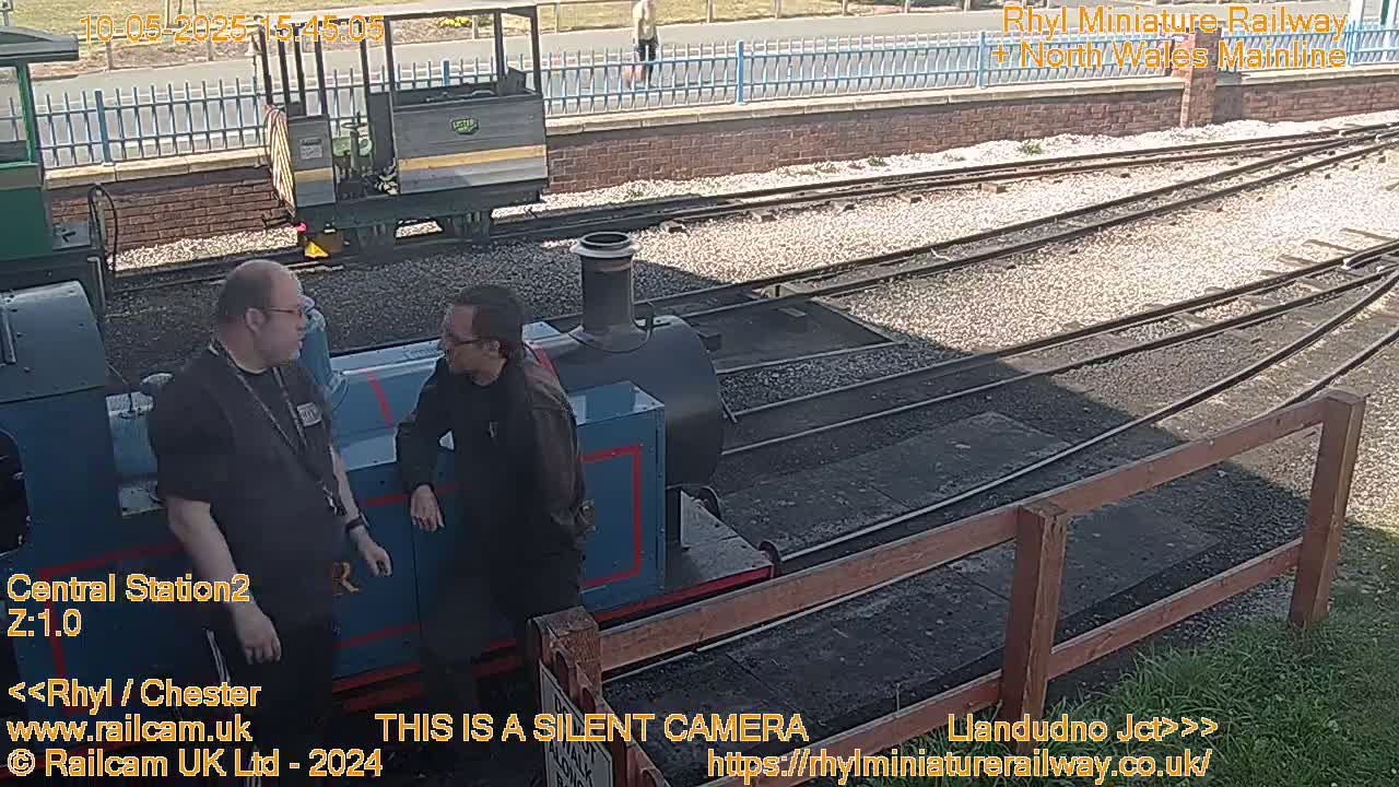 Two men stand beside a small blue train on a sunny day at a railway station.