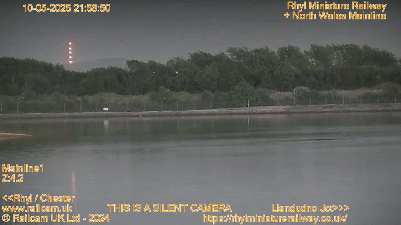 A dark, calm body of water reflects a line of trees under a dusky sky, with several lights visible in the distance.