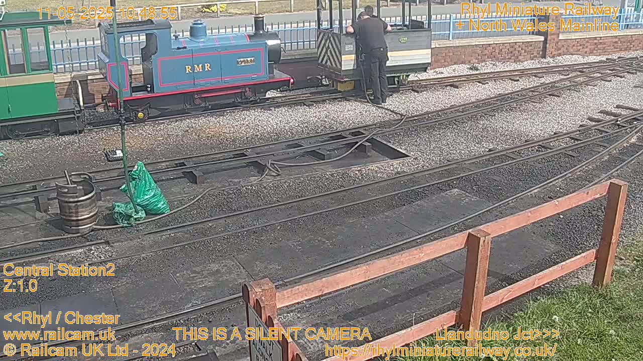 A person is tending to a small, blue train parked on a railway track next to a larger green train, on a sunny day.