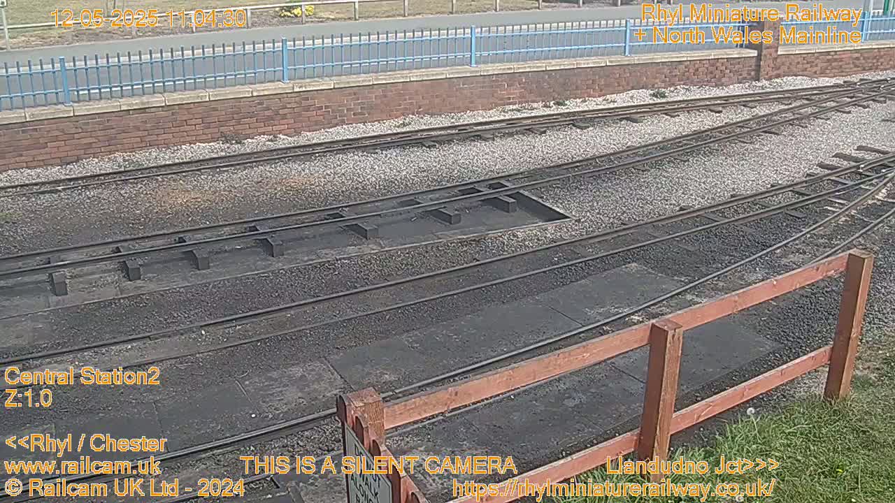 A section of railway tracks with a gravel bed, bordered by a low wooden fence under a partly cloudy sky.