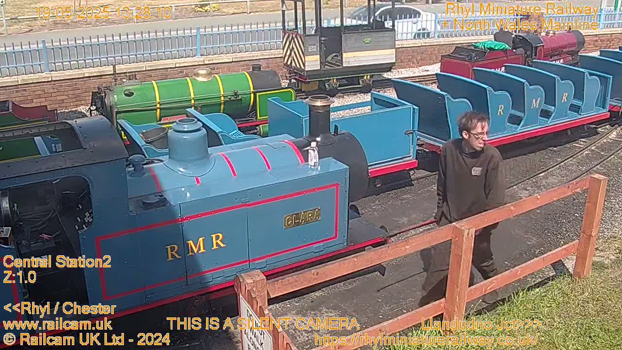 A man stands near a miniature railway on a sunny day, several small locomotives and passenger cars are visible.