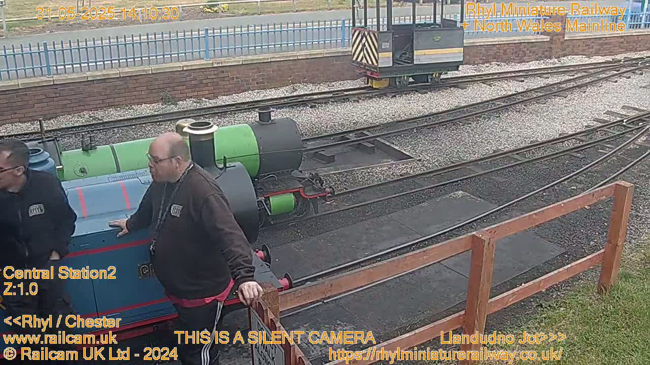 Two men stand near miniature railway trains on a sunny day.