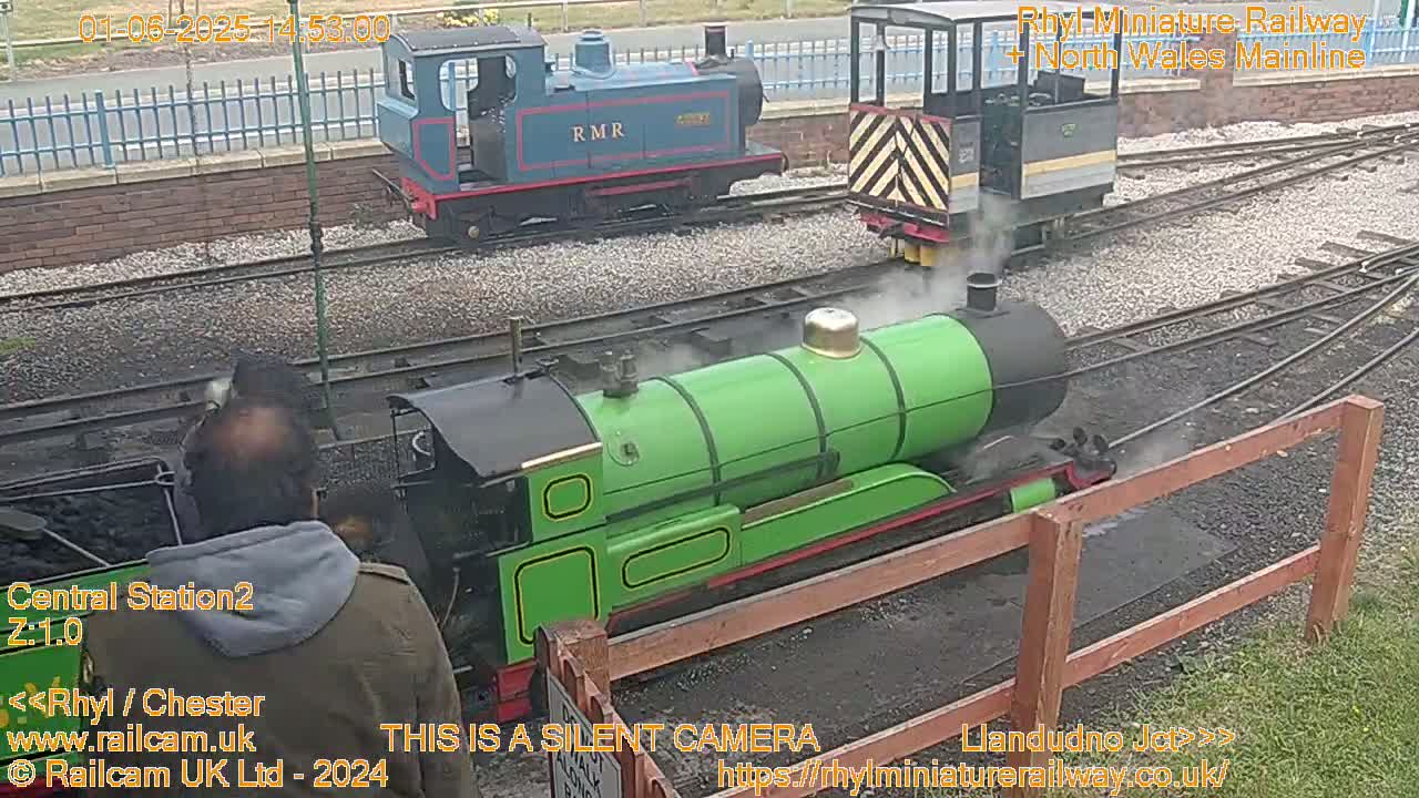 Three miniature train engines, one green, one blue, and one gray, are visible on a railway track in an outdoor setting.