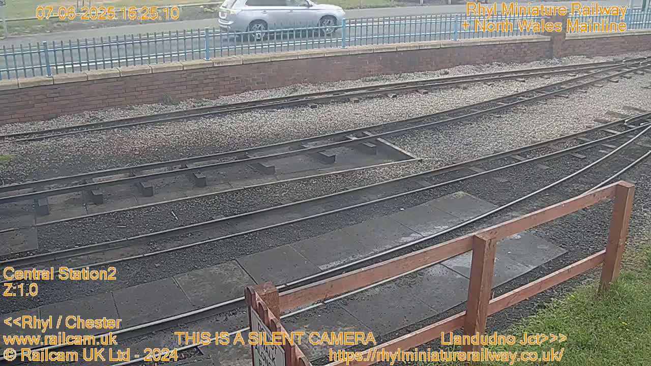A view of railway tracks and a gravel-filled area next to a brick wall, with a silver car visible in the background, under overcast conditions.