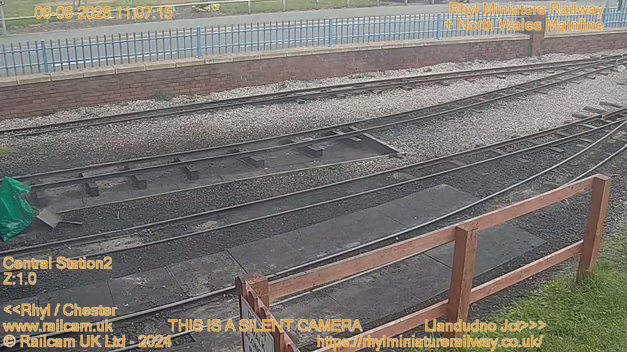 A railway switching yard with multiple tracks and a gravel surface is shown on an overcast day.