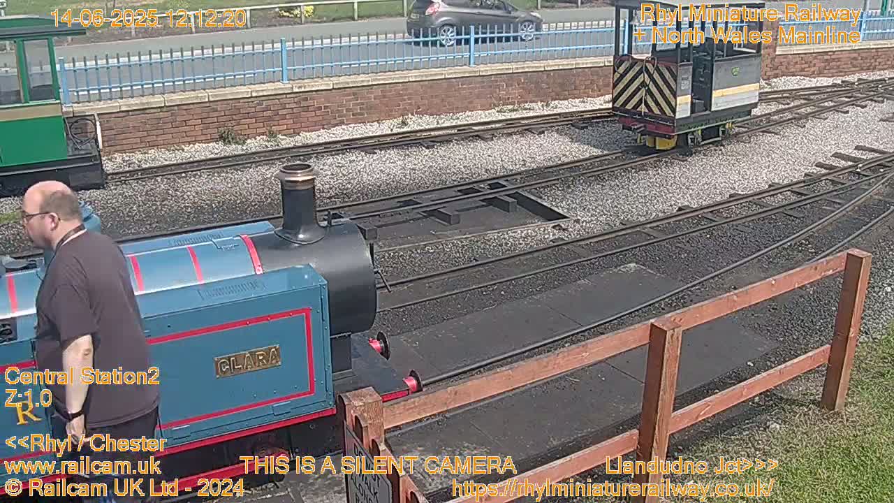 A blue and black miniature train labeled "CLARA" is on a railway track next to a man, with another smaller train visible further down the track on a sunny day.