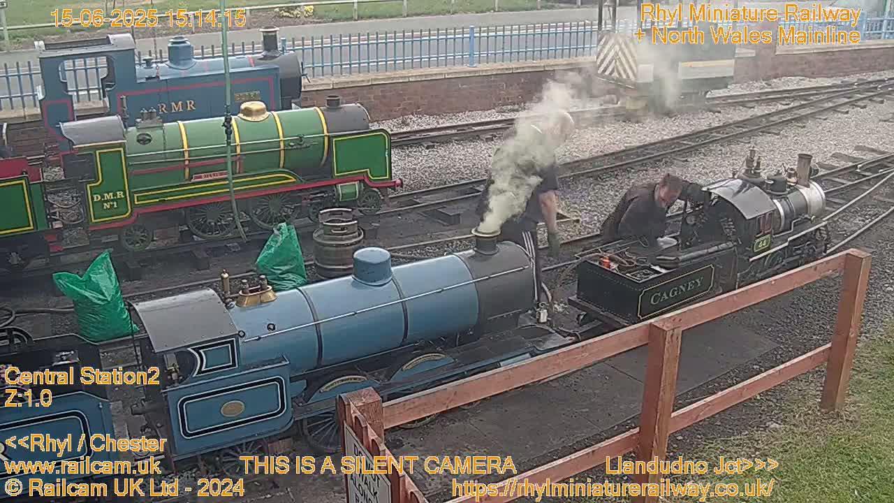 Three miniature locomotives are at a train station, with several people working around them on an overcast day.