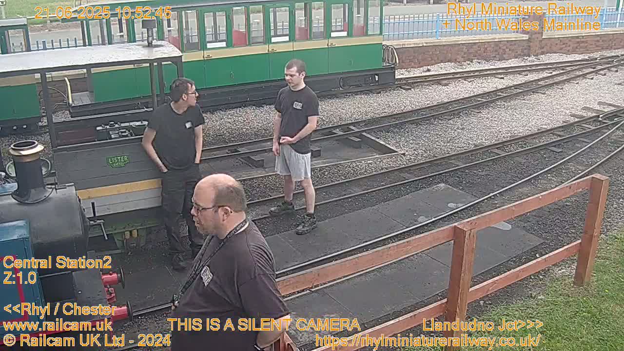 Three men stand near a small railway engine and train tracks on a partly sunny day.