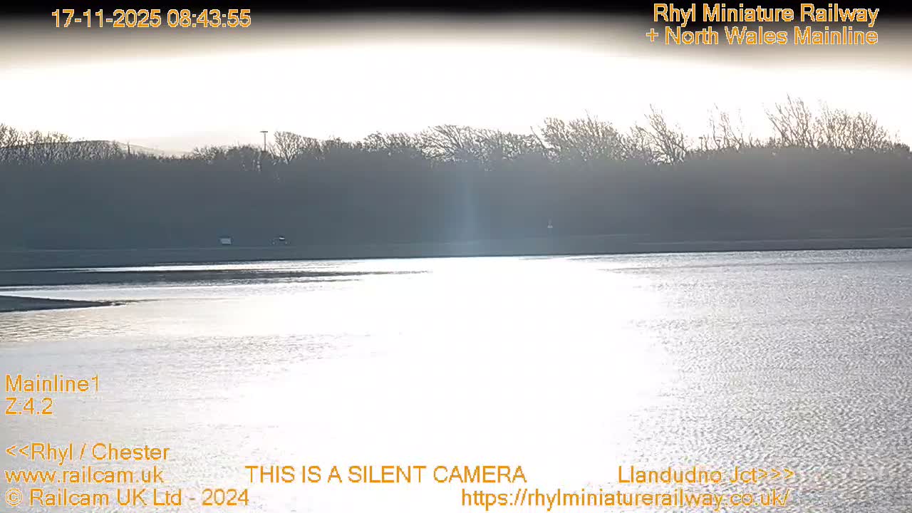 A wide expanse of water gleams intensely with the glare of a low sun under a bright, hazy sky, silhouetting a distant shore lined with bare trees.