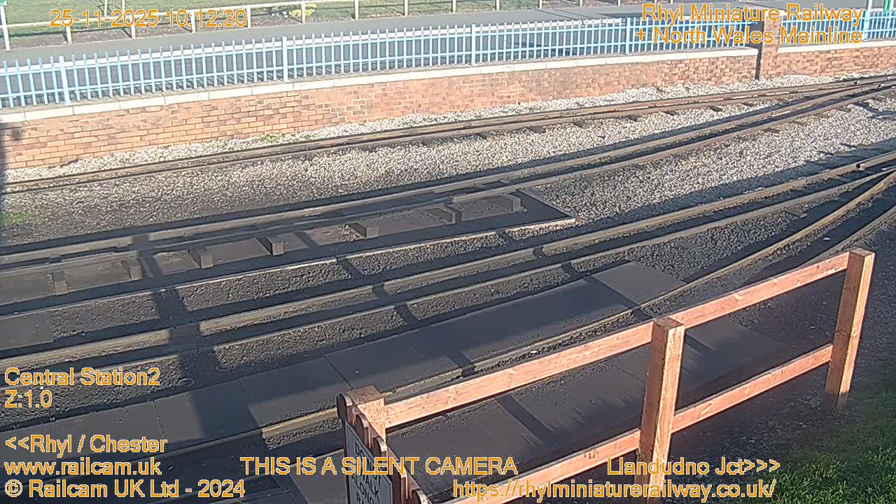 Multiple train tracks, some with switches, run through a sunny outdoor scene bordered by a brick wall, a light blue fence, and a wooden railing in the foreground.