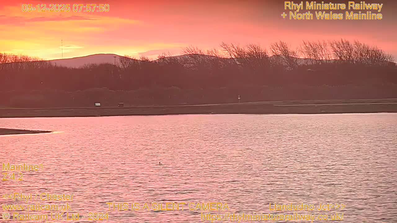 A body of water reflects the vivid orange and red hues of a clear sunrise, with dark land and hills silhouetted against the colorful sky.