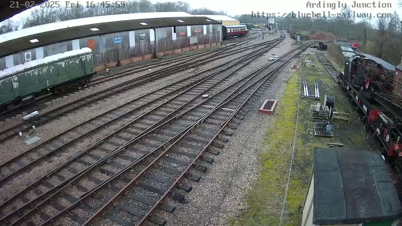 A railway yard with multiple train tracks, several railway cars, and a long building in the background, under an overcast sky.