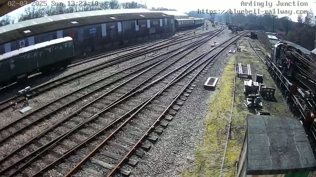 An overhead view of a railway yard on a sunny day shows several train cars and multiple sets of tracks converging near a long building.
