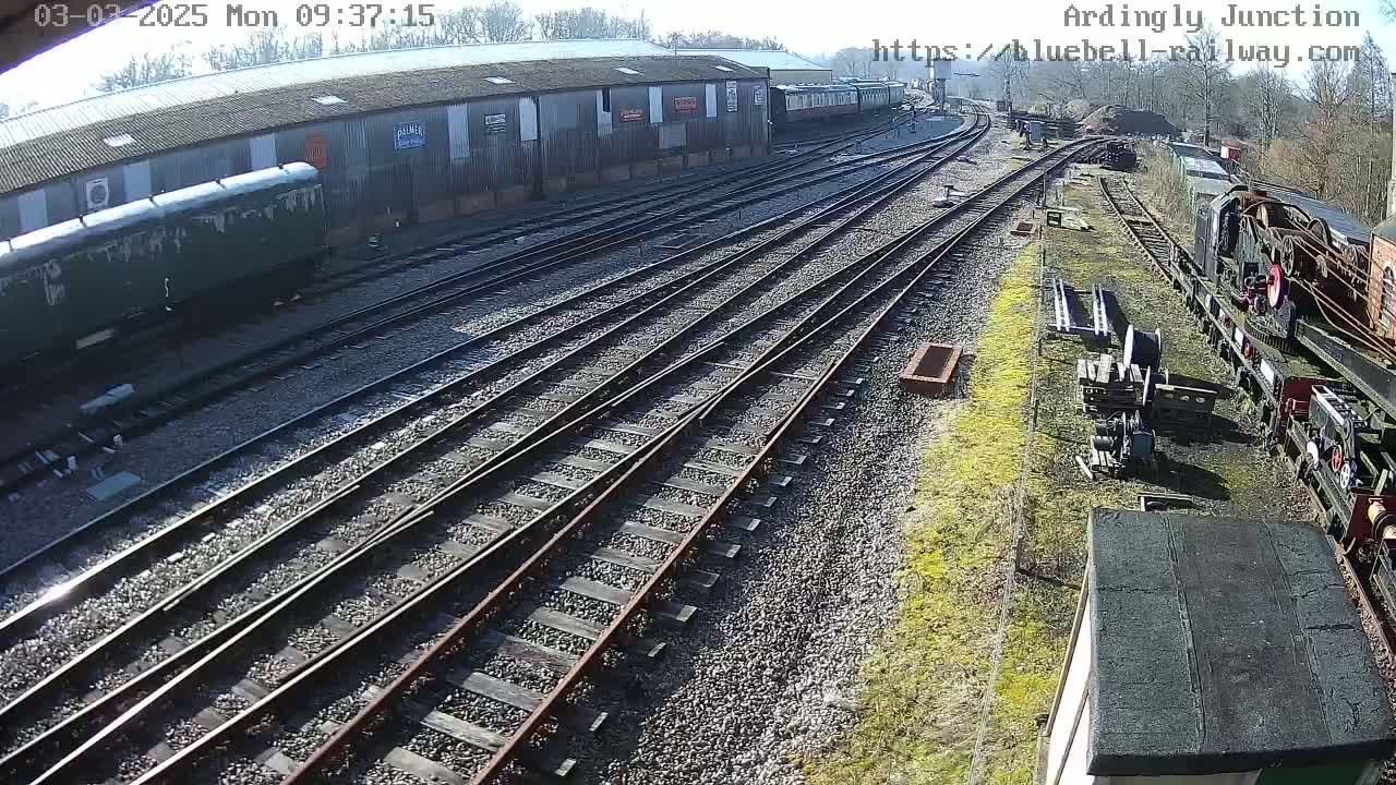A sunny day at a railway yard shows multiple train tracks extending from a large building, with several railway cars and equipment visible.