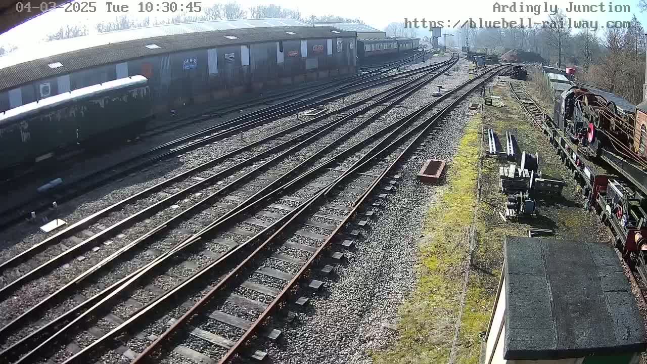 A sunny day at a railway yard shows multiple train tracks converging, with several railway cars and locomotives visible, some of which appear to be in disrepair.