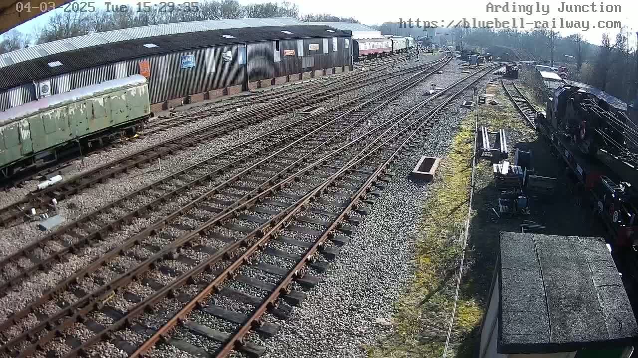 A sunny day at a railway yard shows multiple train tracks branching out from a large shed with several railway carriages and a steam locomotive visible.