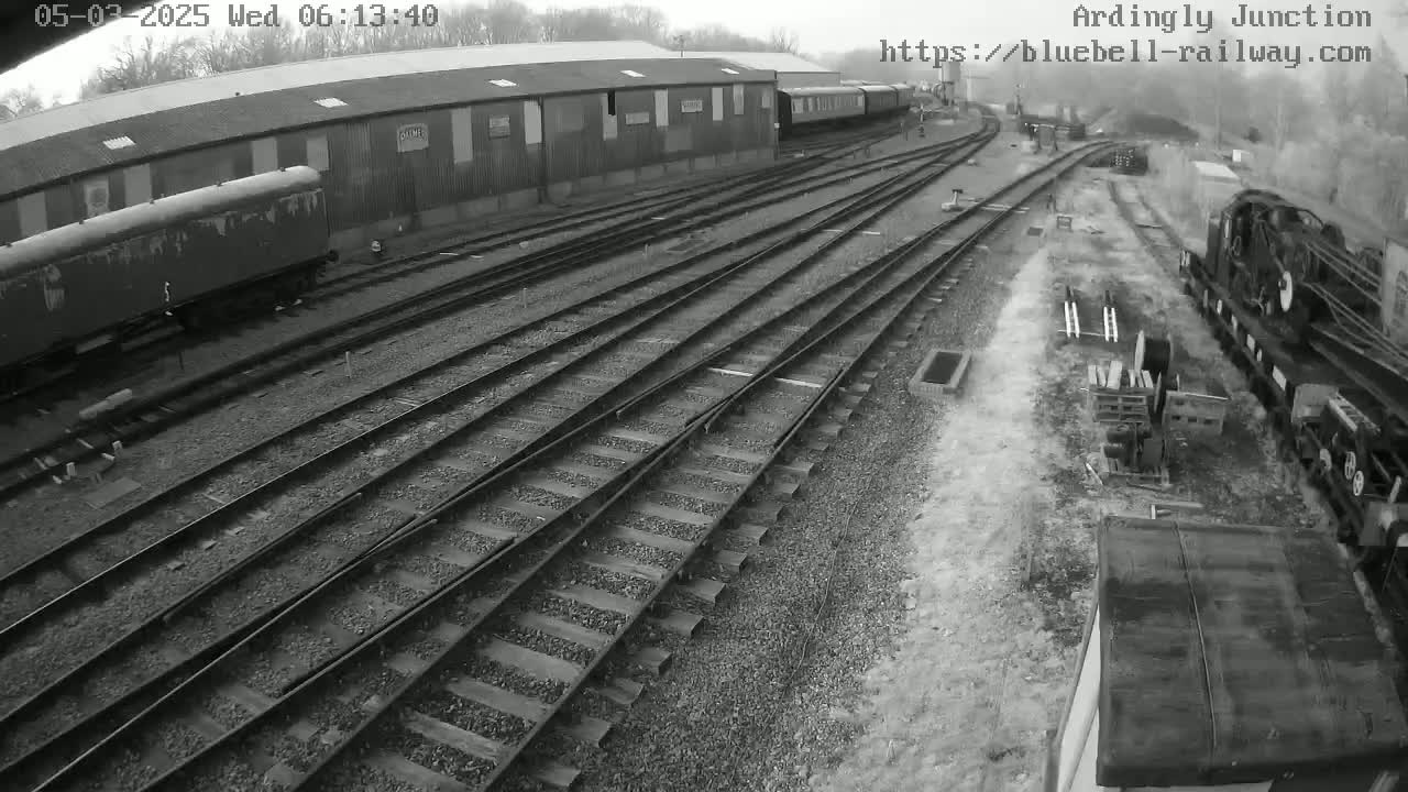 A railway yard with multiple train tracks, several railway cars, and a large building in the background under an overcast sky.