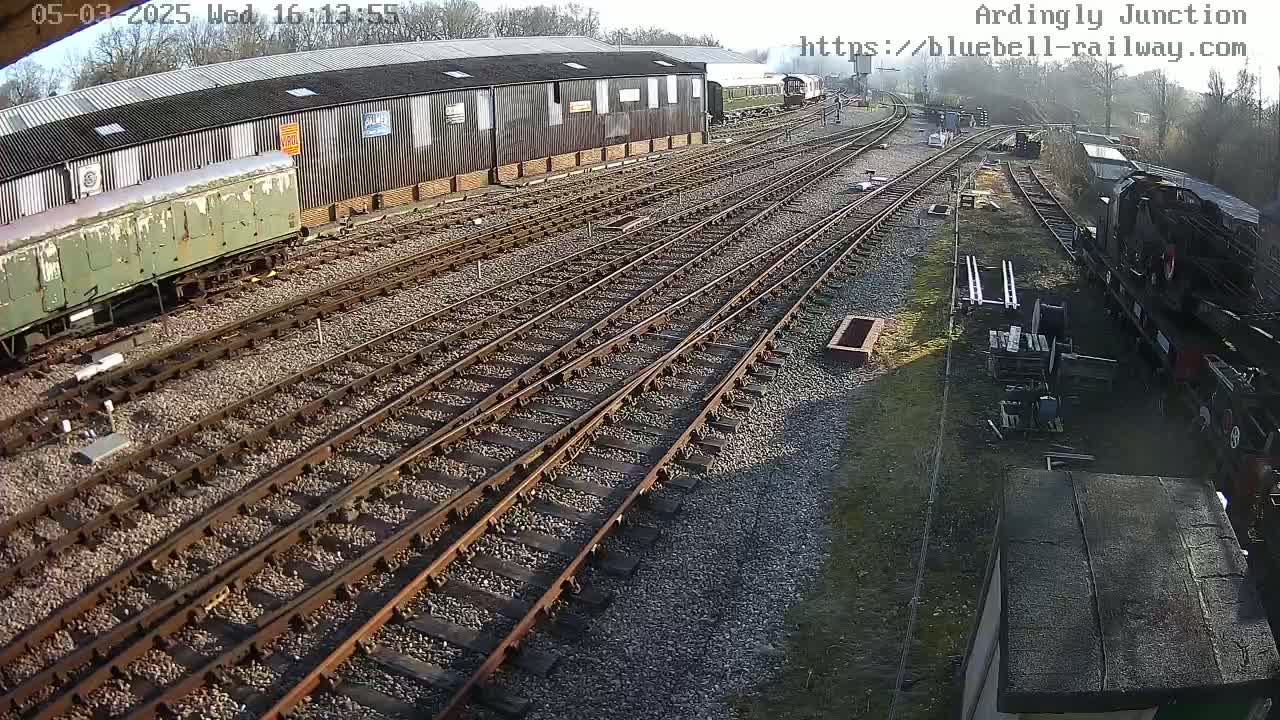 A sunny day at a railway yard shows numerous train tracks intersecting, with several railway cars and locomotives visible.