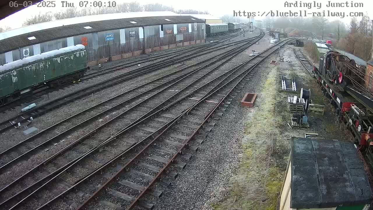 An overcast day at a railway yard shows numerous train tracks, railway cars, and a long building.