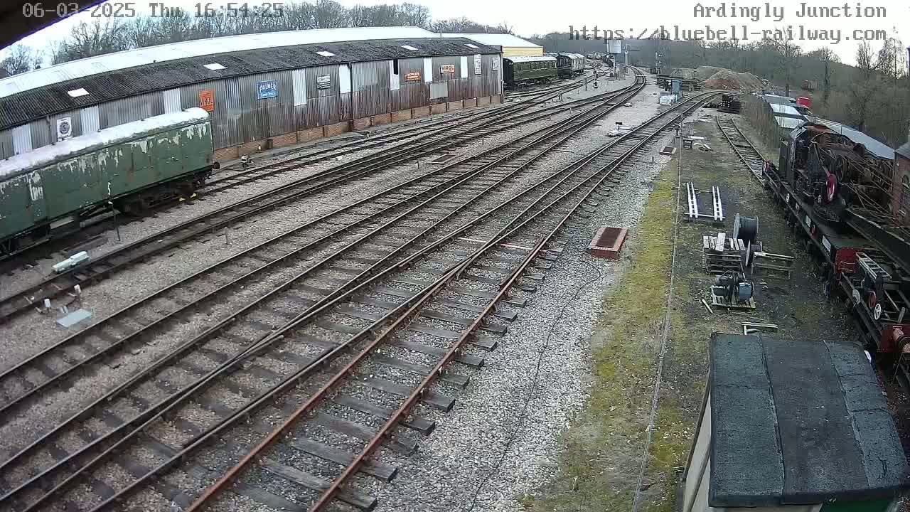 An overcast day reveals a railway yard with multiple train cars, tracks, and a long shed.
