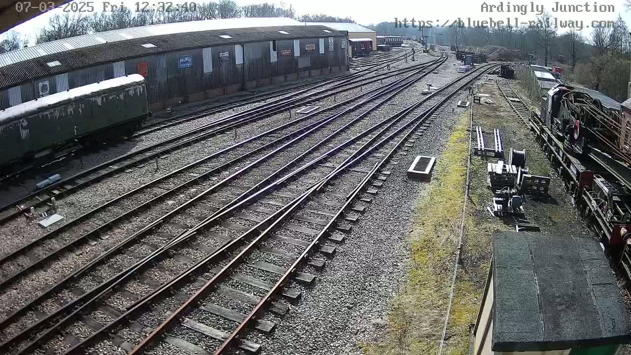 A sunny day at a railway yard shows multiple train tracks converging, with railway cars and a building in the background.