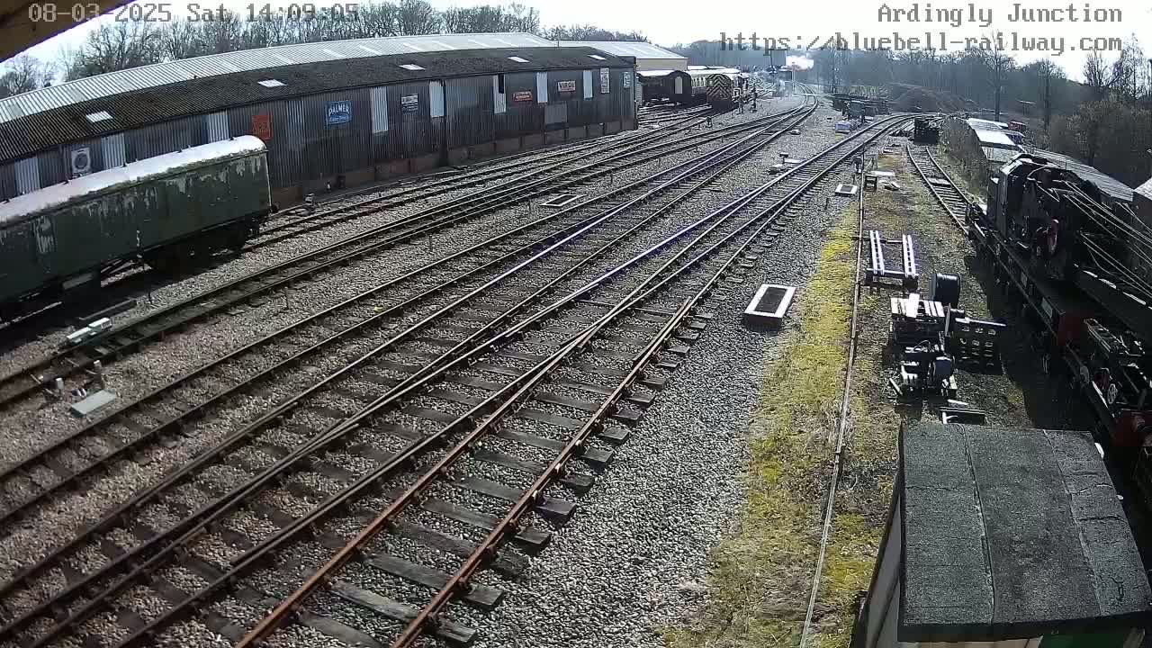 A sunny day at a railway yard shows multiple train tracks branching out from a long building with several rail cars and locomotives parked nearby.