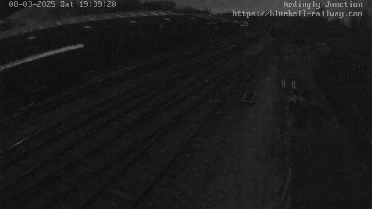 A nighttime, low-light aerial view shows a dark, possibly paved, area with parallel lines, possibly train tracks, extending into the distance.