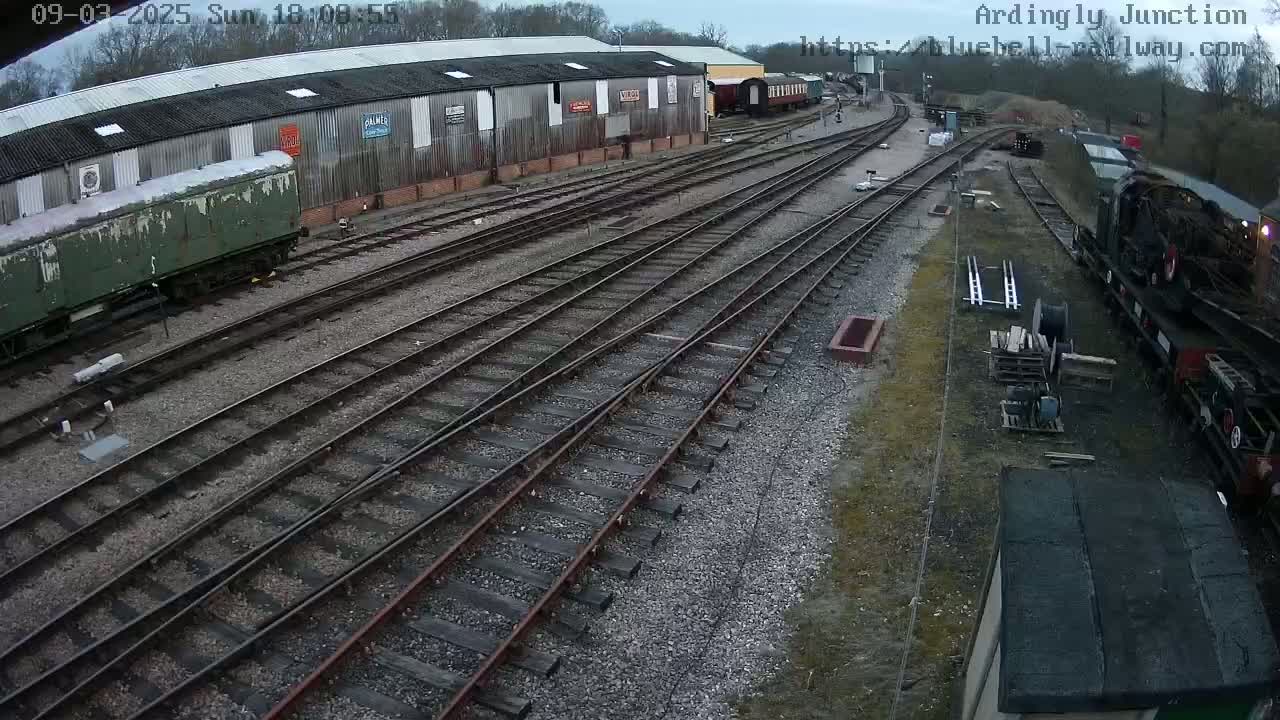 An overcast day reveals a railway yard with multiple train cars and tracks next to a long warehouse.