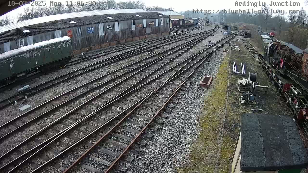 An overcast day at a railway yard shows multiple train tracks branching out from a long, low building with railway cars and locomotives parked nearby.