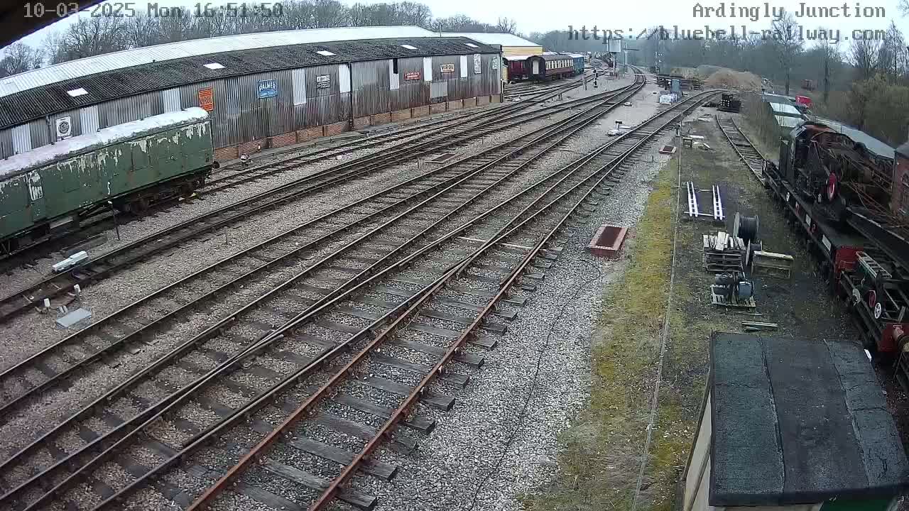 An overcast day shows a railway yard with multiple train cars, railway tracks, and a long building.