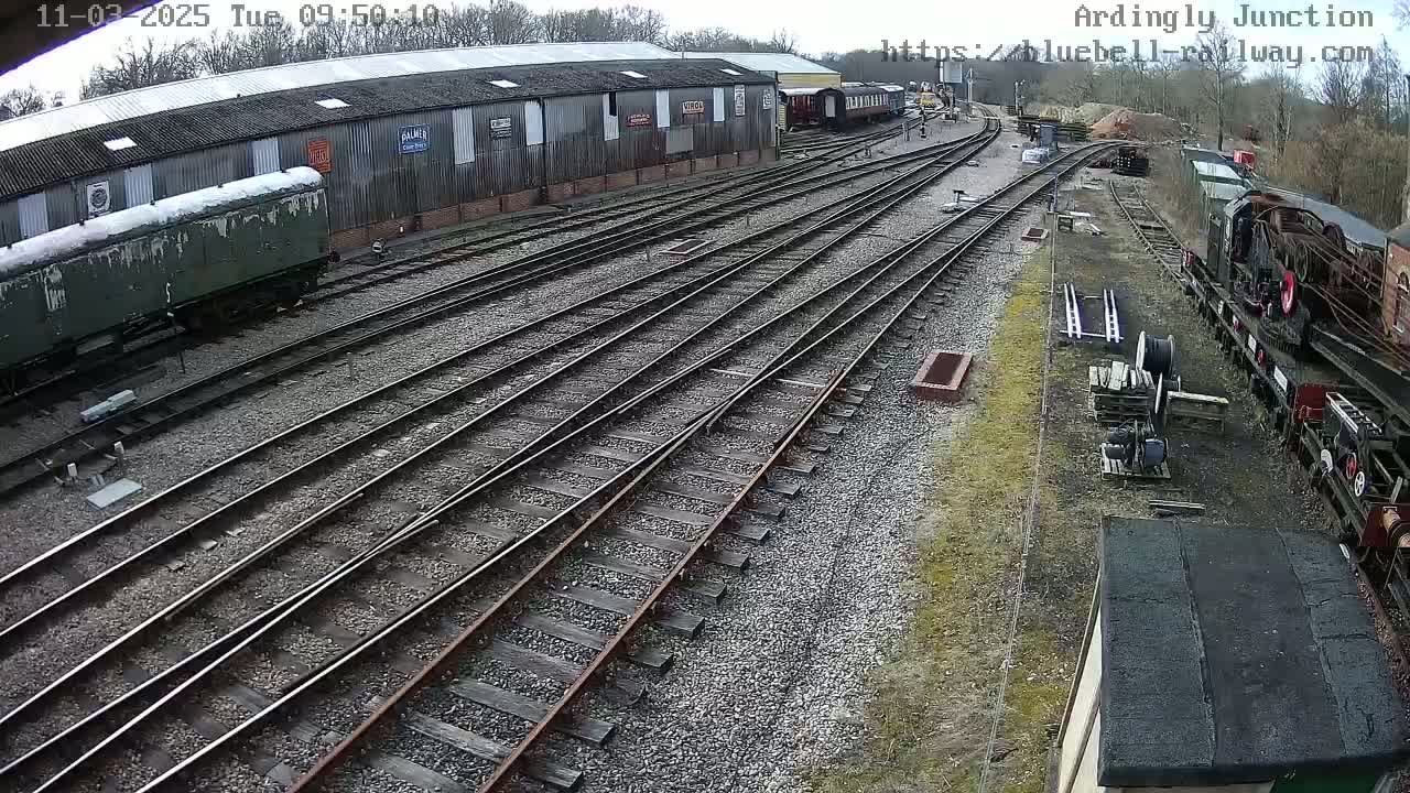 An overhead view shows a railway yard with multiple train tracks, several railway cars, and a long shed under a mostly clear sky.