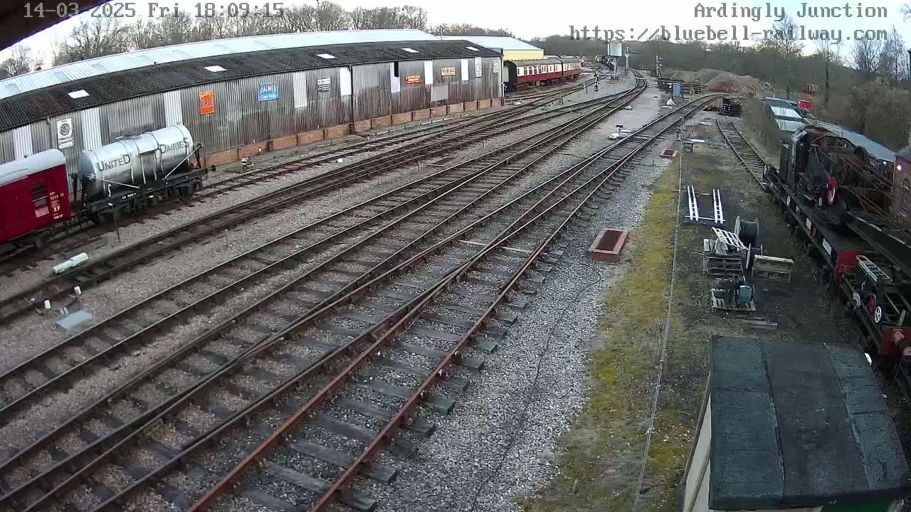 A railway yard with multiple train cars, rail tracks, and a long building is seen on an overcast day.