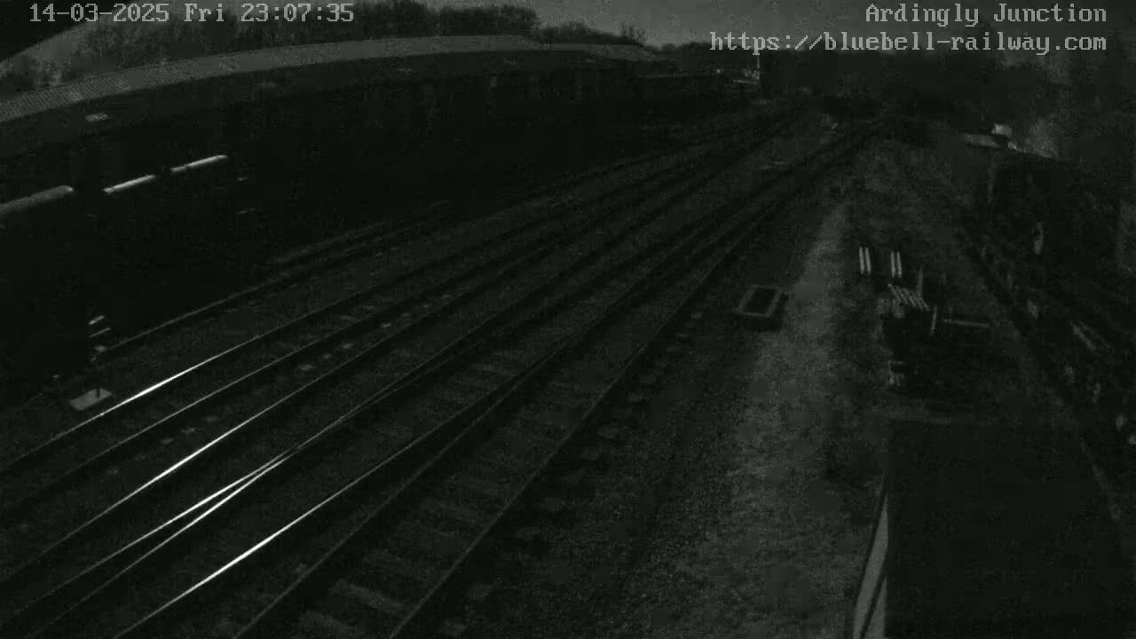 A nighttime, overhead view shows multiple sets of train tracks extending into the distance, with a few dark train cars visible in the left foreground.