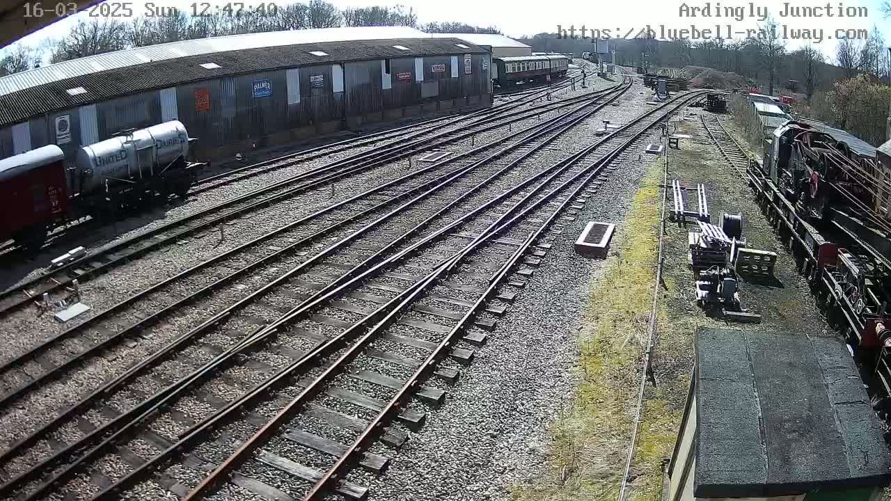 A sunny day at a railway junction shows multiple train tracks branching off from a main line, with various railway cars, including a tank car and a locomotive, parked along the side of a large building.