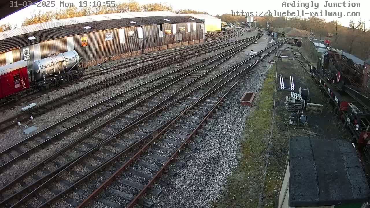 A railway yard with multiple train tracks, several railway cars, and a large industrial building under a daytime sky.