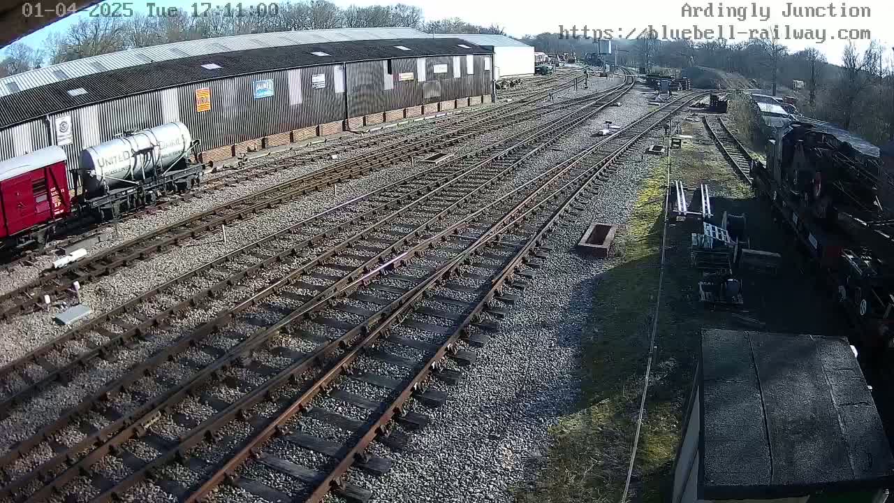 A sunny day at a railway yard shows multiple train tracks branching off from a large building, with several railcars and locomotives present.